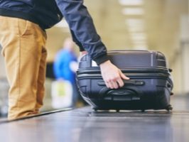 A person reaching for a black suitcase on a luggage carousel at an airport, with blurred figures in the background.