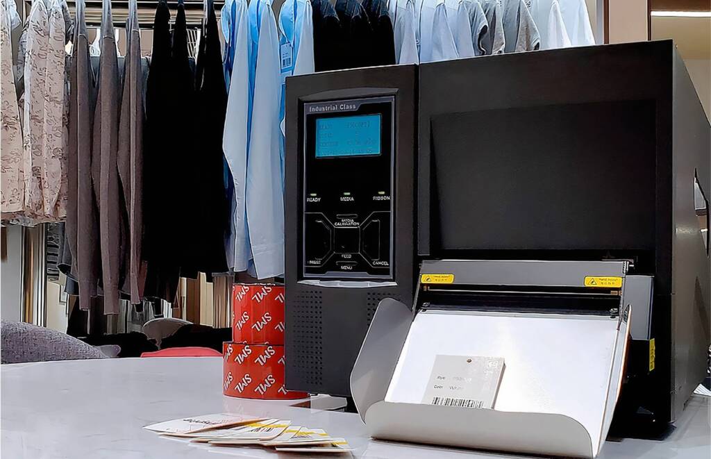 A close-up of an industrial label printer positioned on a countertop, with freshly printed labels in front. In the background, neatly hung clothing items can be seen, indicating a textile or fashion setting. The printer features a digital display and a sleek design.