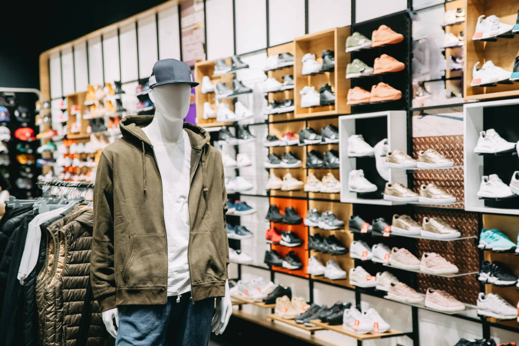 A mannequin dressed in a casual outfit, including a hoodie and jeans, stands in front of a wall filled with various sneaker styles. The vibrant display showcases a wide range of footwear options, creating an engaging shopping environment.
