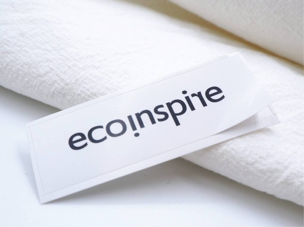 A close-up of a clothing label featuring the "eco.inspire" logo on a white background. The label is partially tucked under a soft fabric, emphasizing its connection to sustainable fashion.