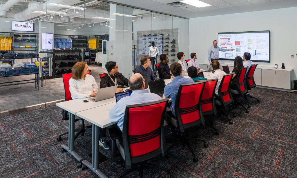 A group of people attending a business presentation in a conference room. The presenter is standing at a large screen displaying slides, while the attendees are seated at a long table with laptops and notebooks.